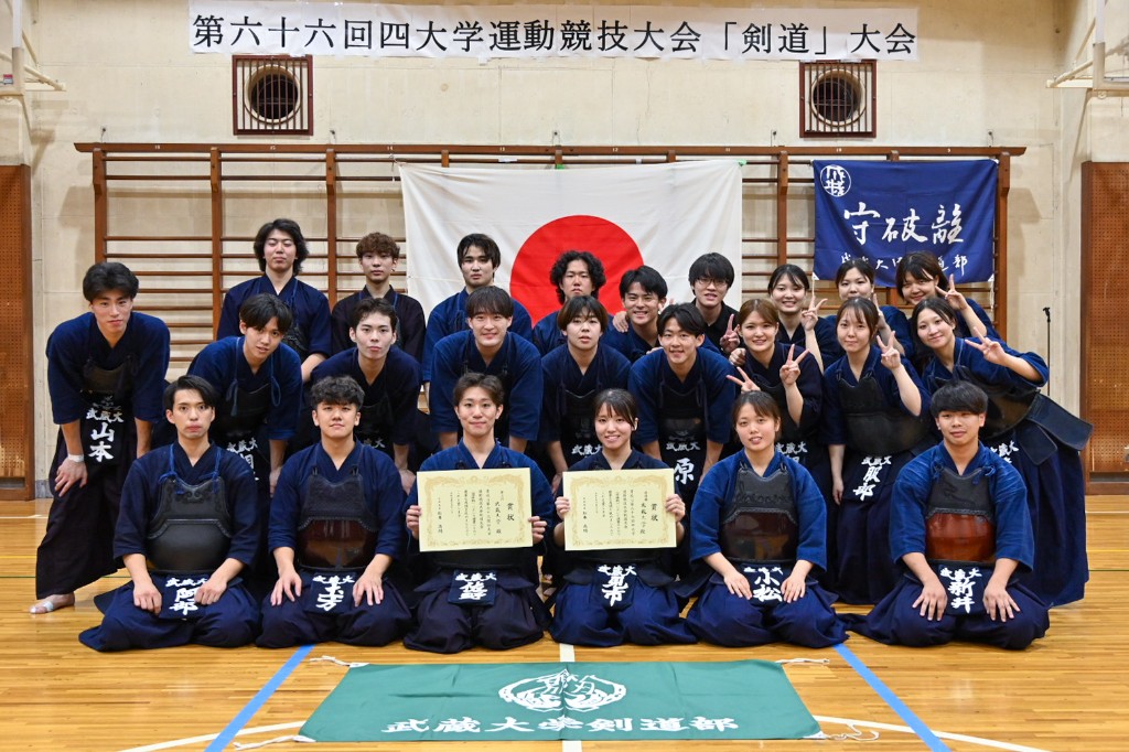 第六十六回四大学運動競技大会剣道の記念撮影。武蔵大学剣道部の部員が道場内で集合し、前列では表彰状を掲げる。背景に大会の横断幕と守破離・武蔵大学剣道部の垂れ幕、前方に部旗がある。