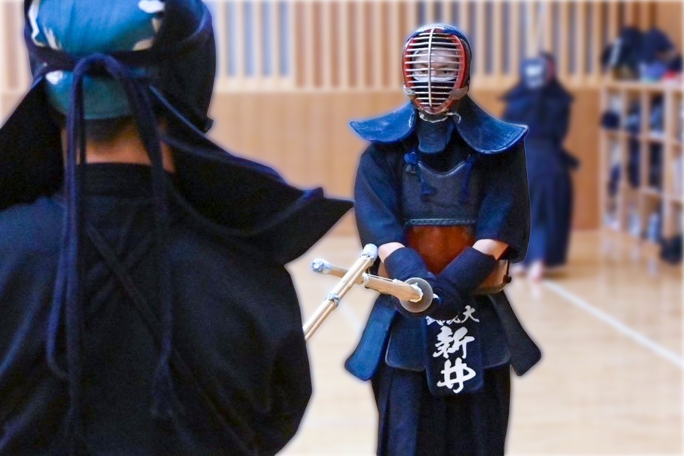 武蔵大学剣道部の道場で、防具姿の部員が中段の構えで竹刀を交え稽古する様子。ゼッケンに武蔵大の文字が見える。