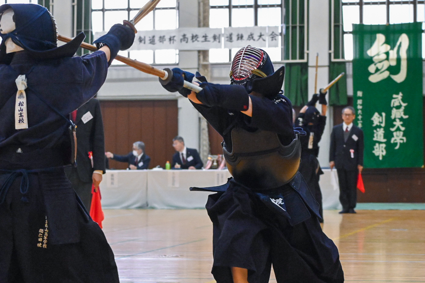 武蔵大学剣道部杯高校生剣道錬成大会の試合で竹刀を交える選手たち。背景に大会名横断幕と武藏大學剣道部の旗。